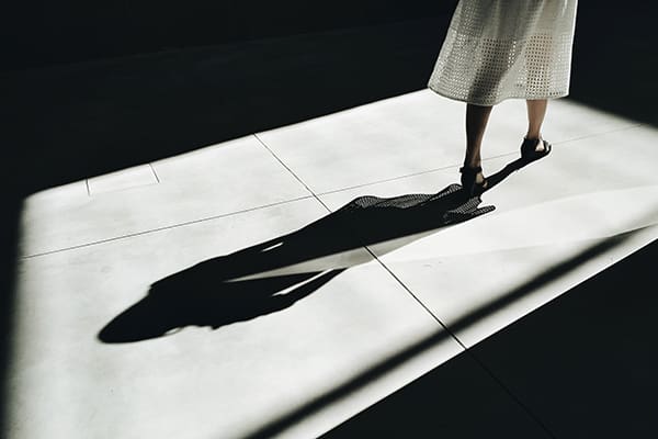 A woman in white dress standing on sidewalk with shadow.